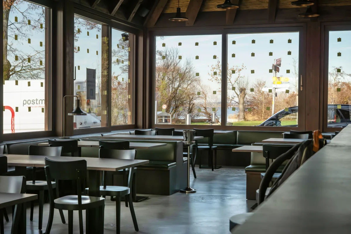 Im Innern kontrastieren ein heller Betonboden und dunkle Holzelemente. Innenansicht des Seerestaurants «im Schilf»: Dunkle Holzmöbel auf einem hellen Betonboden und grosse Fensterflächen.